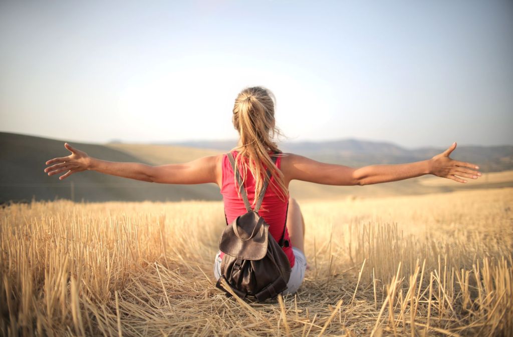 A woman sitting in a harvested field with her arms outstretched, enjoying the scenery under a clear sky.