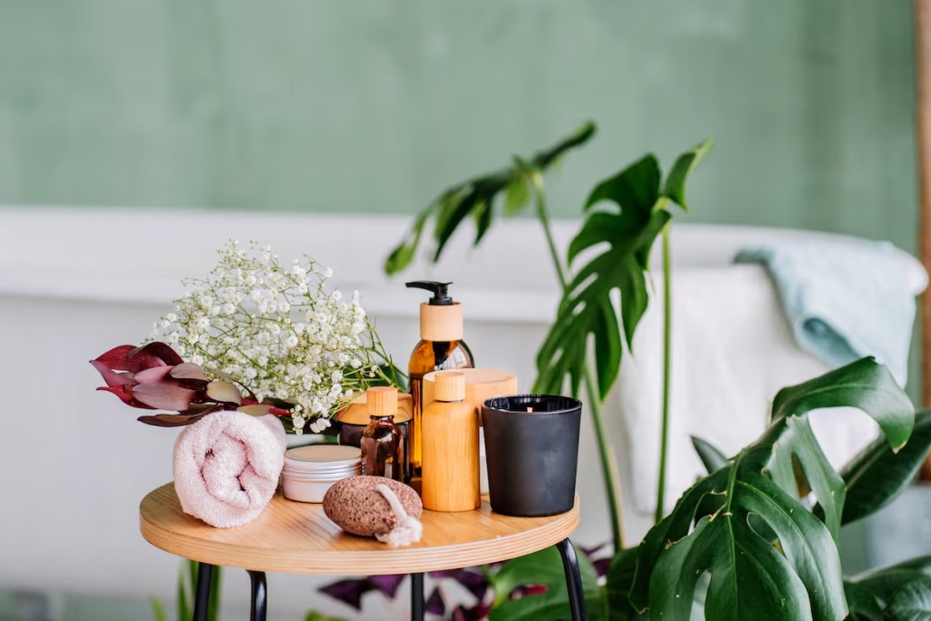 A wooden table displaying various skincare products, including bottles and jars, alongside a bouquet of white flowers and a rolled towel, set against a green wall and surrounded by indoor plants.
